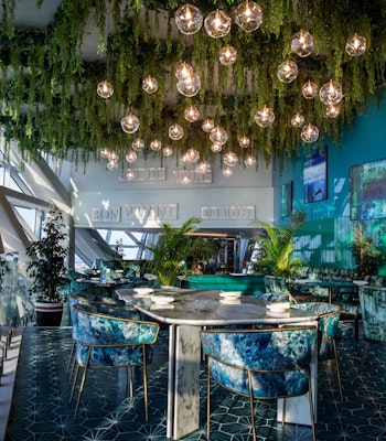 Andaz Capital Gate Abu Dhabi A stylish dining area with hanging lights and greenery, featuring elegant tables and chairs in vibrant colors.