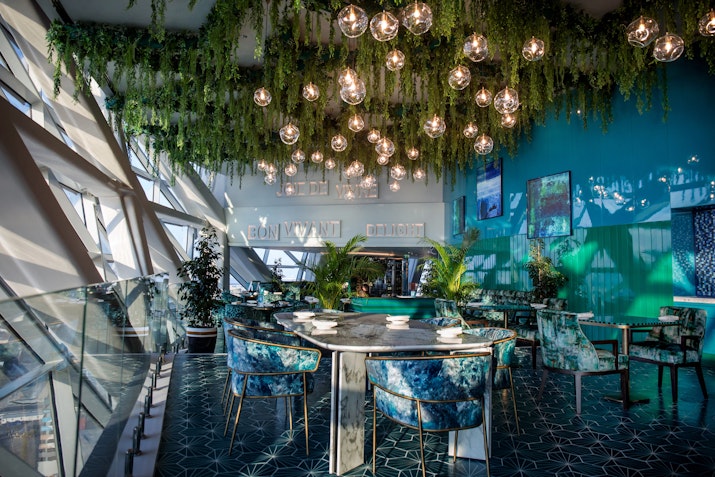 A stylish dining area with hanging lights and greenery, featuring elegant tables and chairs in vibrant colors.