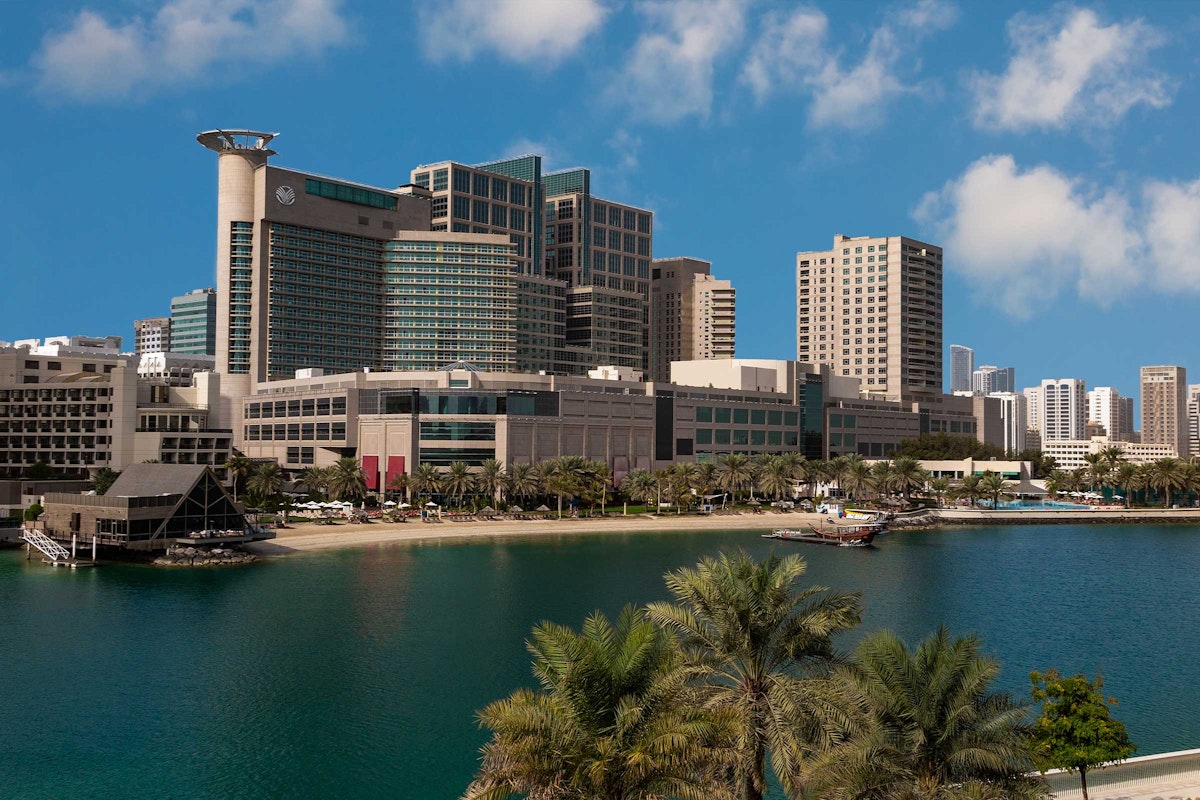 A modern hotel complex overlooking a waterfront with palm trees, surrounded by city skyscrapers and a bright sky.