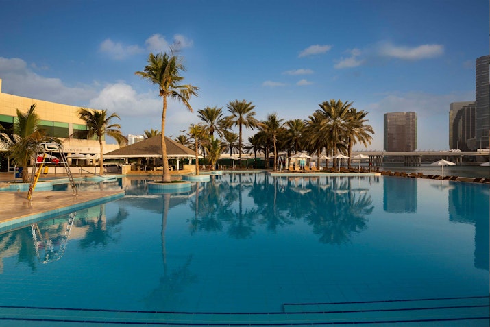 A tranquil outdoor pool with clear blue water, surrounded by palm trees and modern buildings under a bright sky.