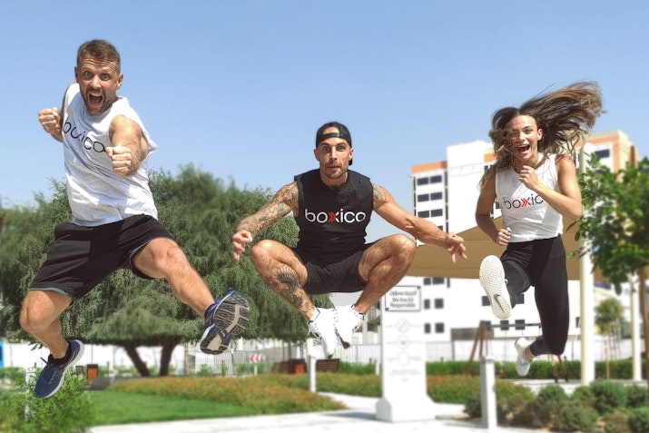 Three energetic fitness trainers from Body-Co leaping in mid-air outdoors, displaying excitement and enthusiasm. They are dressed in athletic gear, with a clear blue sky and greenery in the background, creating a lively and motivational scene