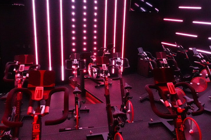 A dark cycling studio with rows of stationary bikes illuminated by intense pink and red neon lights. Vertical and horizontal LED light strips create a dynamic atmosphere, enhancing the energetic vibe of the workout space