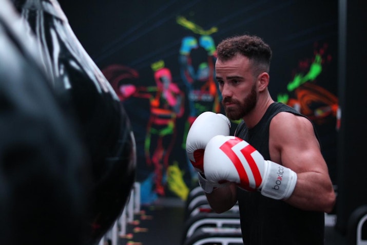 A focused male boxer in a black tank top and white gloves practicing punches on a punching bag in a vibrant gym setting. Neon lights and colorful artwork on the walls create an energetic and intense atmosphere, highlighting his dedication to training.