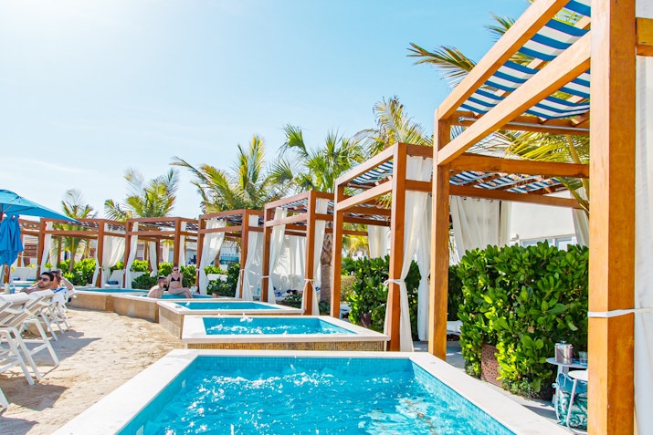 Beachside cabanas with private hot tubs surrounded by lush greenery and palm trees under a clear blue sky.