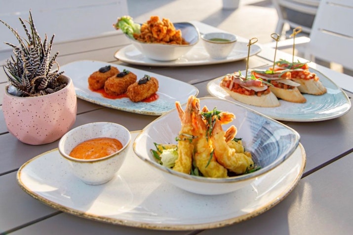 A selection of gourmet appetizers including shrimp, croquettes, and bruschetta, beautifully presented on plates with a small potted plant in the background.