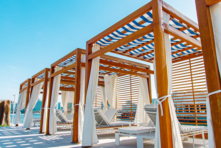 A row of wooden cabanas with white curtains and striped canopies, creating a relaxing beachside atmosphere under a clear blue sky.