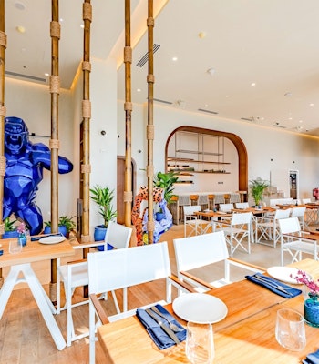 COVEBEACH Dining A modern restaurant interior featuring wooden tables and white chairs, with a large blue sculpture of a figure and greenery in the decor.