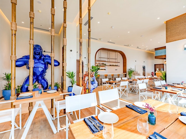 A modern restaurant interior featuring wooden tables and white chairs, with a large blue sculpture of a figure and greenery in the decor.
