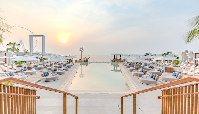 COVEBEACH Dubai A view of a pool area at sunset, with steps leading down to lounge seating and umbrellas, overlooking the water and a distant Ferris wheel.