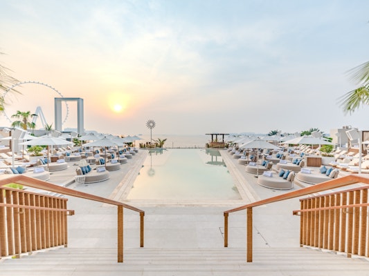 A view of a pool area at sunset, with steps leading down to lounge seating and umbrellas, overlooking the water and a distant Ferris wheel.