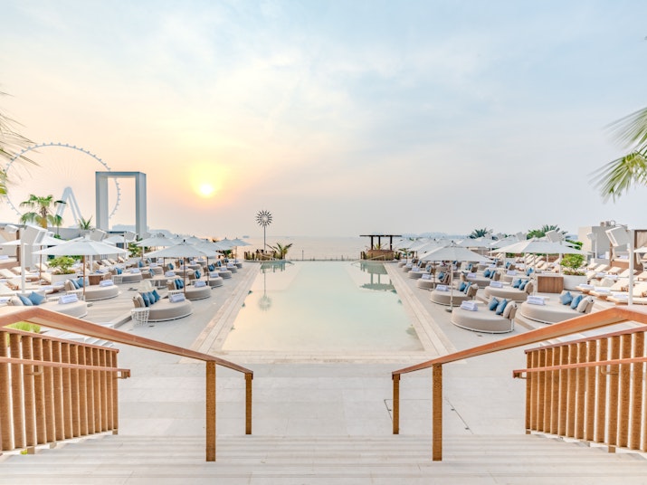 A view of a pool area at sunset, with steps leading down to lounge seating and umbrellas, overlooking the water and a distant Ferris wheel.