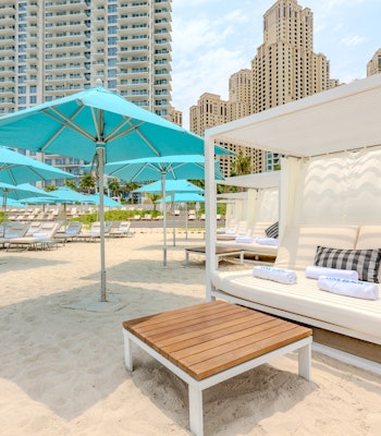 COVEBEACH Dubai A beach cabana with white cushions and a wooden table, surrounded by turquoise umbrellas and lounge chairs, with tall buildings in the background.