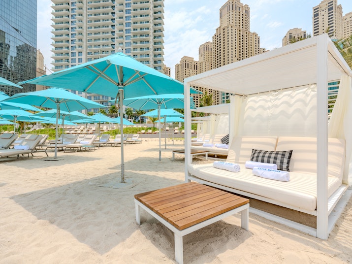 A beach cabana with white cushions and a wooden table, surrounded by turquoise umbrellas and lounge chairs, with tall buildings in the background.