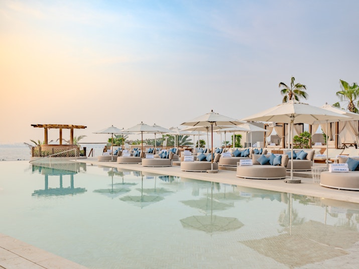 Lounge chairs and umbrellas beside a tranquil pool at sunset, with a backdrop of the ocean and a wooden gazebo.