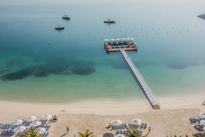 Aerial view of a tranquil beach with a wooden pier extending into the calm water, surrounded by boats and beach umbrellas.