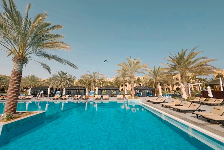 A swimming pool surrounded by palm trees and sun loungers under a clear sky.