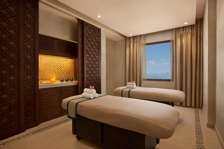 A serene spa room featuring two massage tables with towels and decorative accents, large window offering a view, and a soothing atmosphere.