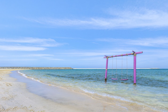 A colorful pink swing set on a sandy beach beside calm turquoise waters, under a clear blue sky.