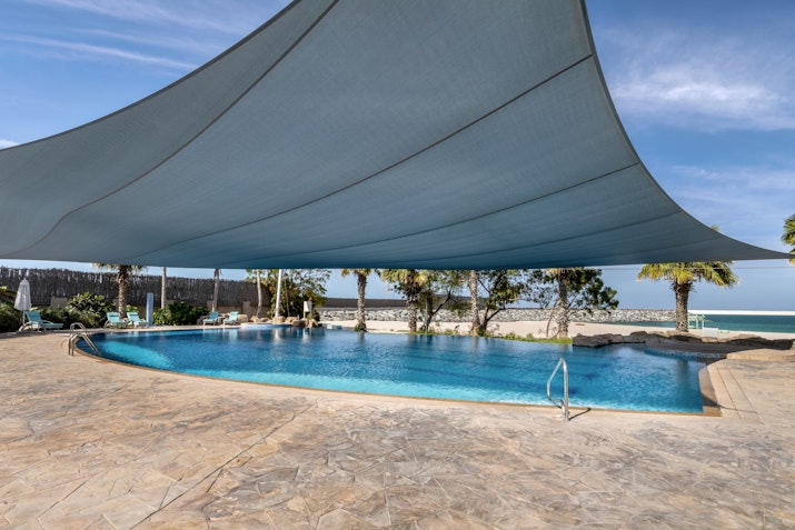 A tranquil swimming pool shaded by a large blue canopy, surrounded by palm trees and a view of the beach in the background, under a clear sky.