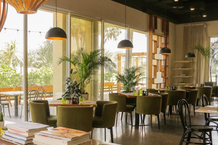 A cozy café interior featuring green upholstered chairs, wooden tables, and large windows that allow natural light to fill the space, complemented by decorative plants.