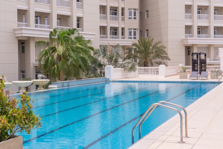 Outdoor swimming pool with clear blue water, surrounded by palm trees and a backdrop of modern architecture. Poolside features include comfortable lounge areas and tropical landscaping, creating a peaceful retreat in the heart of the city.