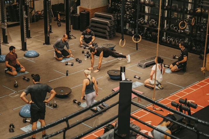 Group fitness class in a gym with participants engaged in various exercises, surrounded by weights, equipment, and ropes. The atmosphere is energetic and focused with a mix of people performing different workout moves on marked mats