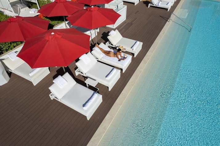Aerial view of a poolside area with red umbrellas shading white loungers and a person relaxing by the pool.