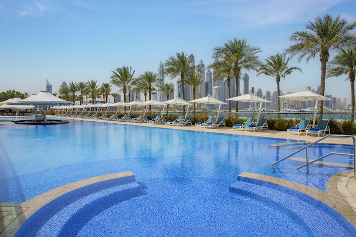 A luxurious pool with sparkling blue water surrounded by palm trees, lounge chairs, and umbrellas, set against a city skyline backdrop.