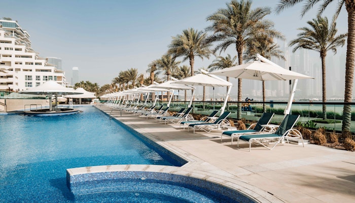 Hilton Dubai Palm Jumeirah A serene pool area featuring blue water, palm trees, and lounge chairs under white umbrellas, with modern buildings in the background.