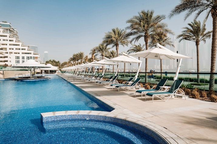 A serene pool area featuring blue water, palm trees, and lounge chairs under white umbrellas, with modern buildings in the background.