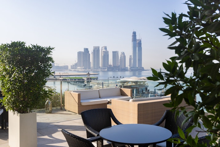 Outdoor seating area with plants and a table, overlooking a city skyline with tall buildings beside the water.