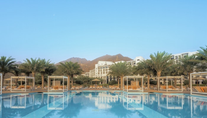 InterContinental Fujairah Resort A stunning hotel pool with cabanas surrounded by palm trees and mountains in the background, under a clear blue sky.