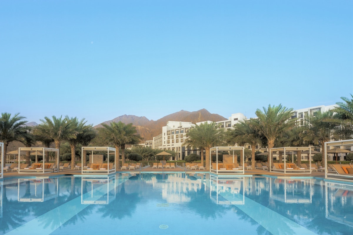 InterContinental Fujairah Resort A stunning hotel pool with cabanas surrounded by palm trees and mountains in the background, under a clear blue sky.