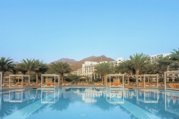A stunning hotel pool with cabanas surrounded by palm trees and mountains in the background, under a clear blue sky.
