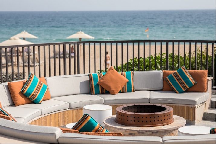 A cozy seating area with colorful cushions, overlooking a beach and the sea, with a fire pit at the center.