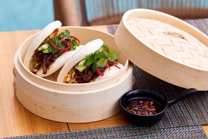 InterContinental Ras Al Khaimah Two steamed buns filled with meat, garnished with cilantro and chili, served in a bamboo steamer with a side of dipping sauce.
