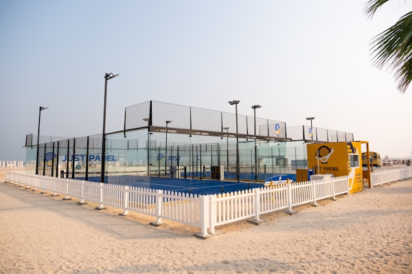 Just Padel Kite Beach Outdoor padel court by Just Padel, situated on a sandy beachfront, surrounded by a white picket fence. The setup includes clear glass walls, vibrant blue court surface, and nearby palm trees, creating a serene beachside sports environment