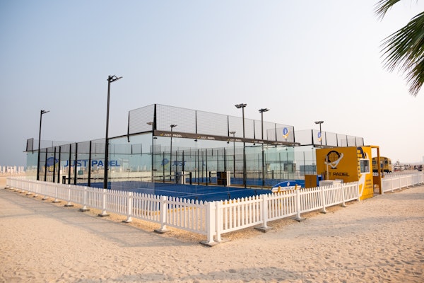 Outdoor padel court by Just Padel, situated on a sandy beachfront, surrounded by a white picket fence. The setup includes clear glass walls, vibrant blue court surface, and nearby palm trees, creating a serene beachside sports environment