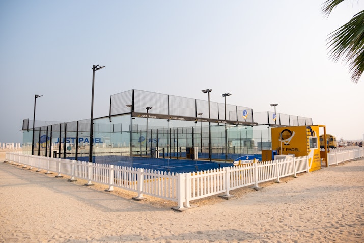 Outdoor padel court by Just Padel, situated on a sandy beachfront, surrounded by a white picket fence. The setup includes clear glass walls, vibrant blue court surface, and nearby palm trees, creating a serene beachside sports environment