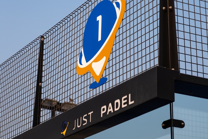 Close-up view of Court No. 1 at Just Padel, featuring a bold blue and orange logo on the mesh fence above the court, with the text 'Just Padel' displayed below. The setup reflects a modern and professional padel court environment.
