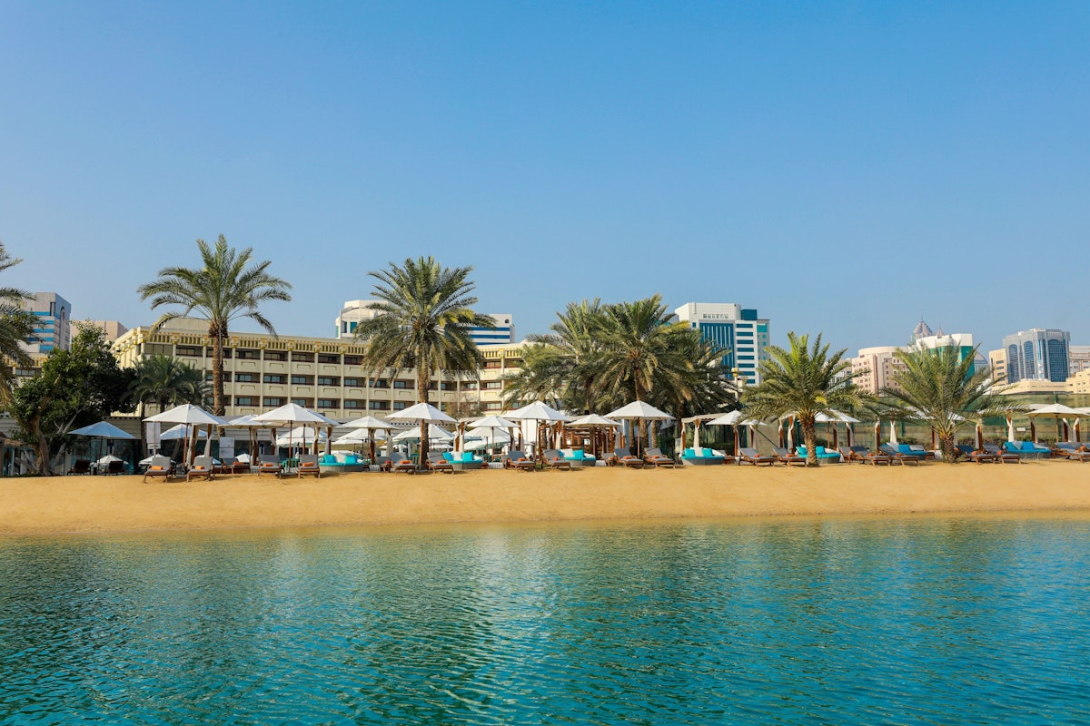 Le Meridien Abu Dhabi A scenic view of a beach with sun loungers and umbrellas, framed by palm trees and a backdrop of modern buildings under a clear blue sky.