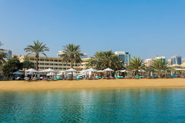 A scenic view of a beach with sun loungers and umbrellas, framed by palm trees and a backdrop of modern buildings under a clear blue sky.