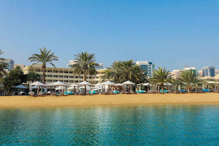 A scenic view of a beach with sun loungers and umbrellas, framed by palm trees and a backdrop of modern buildings under a clear blue sky.