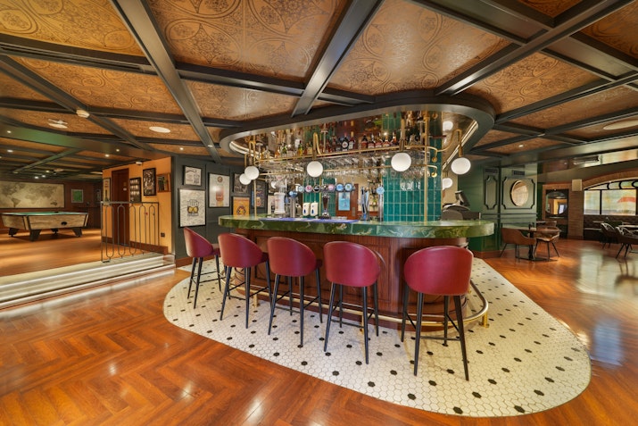 An elegant bar area featuring a curved counter with high chairs, stylish decor, and an array of bottles on display. The warm wooden floors and rich colors create a sophisticated atmosphere.