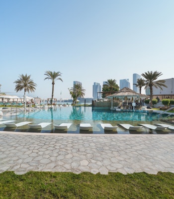 Le Meridien Abu Dhabi A bright pool area surrounded by sun loungers and palm trees, with modern buildings in the background and a clear blue sky.