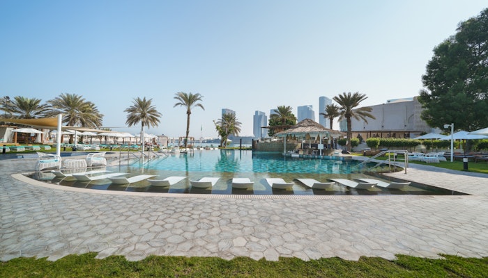 Le Meridien Abu Dhabi A bright pool area surrounded by sun loungers and palm trees, with modern buildings in the background and a clear blue sky.
