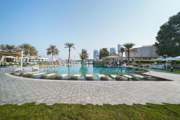 A bright pool area surrounded by sun loungers and palm trees, with modern buildings in the background and a clear blue sky.