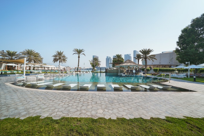 A bright pool area surrounded by sun loungers and palm trees, with modern buildings in the background and a clear blue sky.
