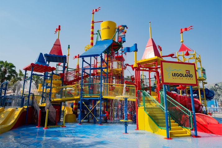 Brightly colored water play structure at LEGOLAND Water Park in Dubai, featuring red, yellow, and blue slides, water cannons, and interactive climbing areas, all under a clear blue sky.