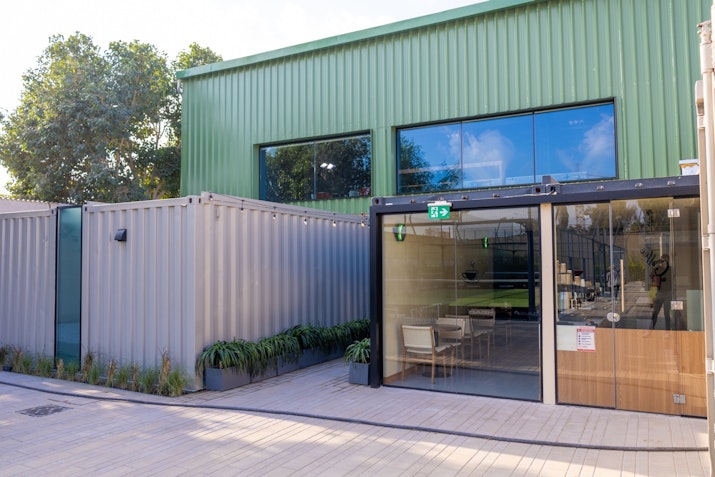 Mantra An exterior view of a clubhouse with a modern industrial design, featuring green metal siding and large, clear glass windows. The entrance has a glass door framed in black with a green emergency exit sign above. Adjacent to the building are corrugated metal walls and neatly arranged potted plants, adding greenery to the minimalist and functional aesthetic of the structure